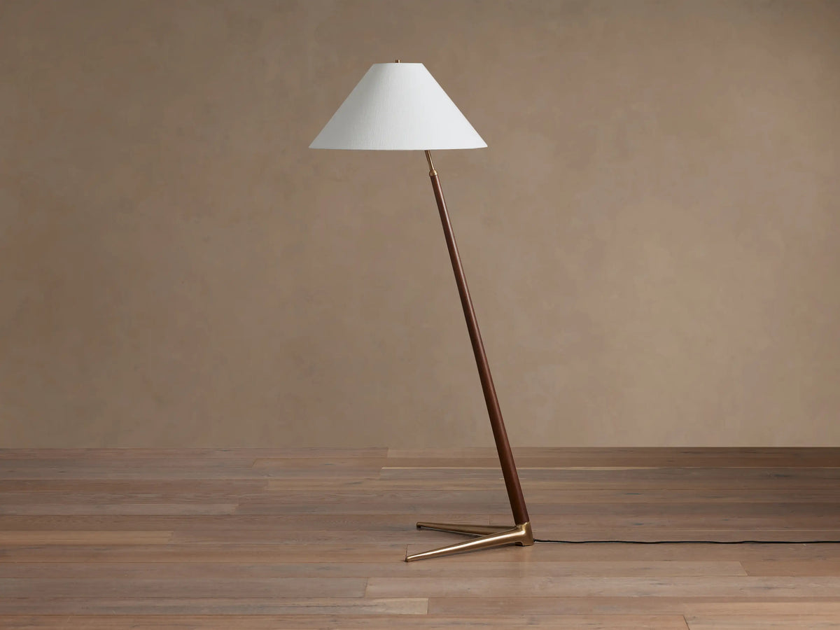 Angled Edric Floor Lamp with walnut wood pole, white conical linen shade, and brass tripod base on a wooden floor.