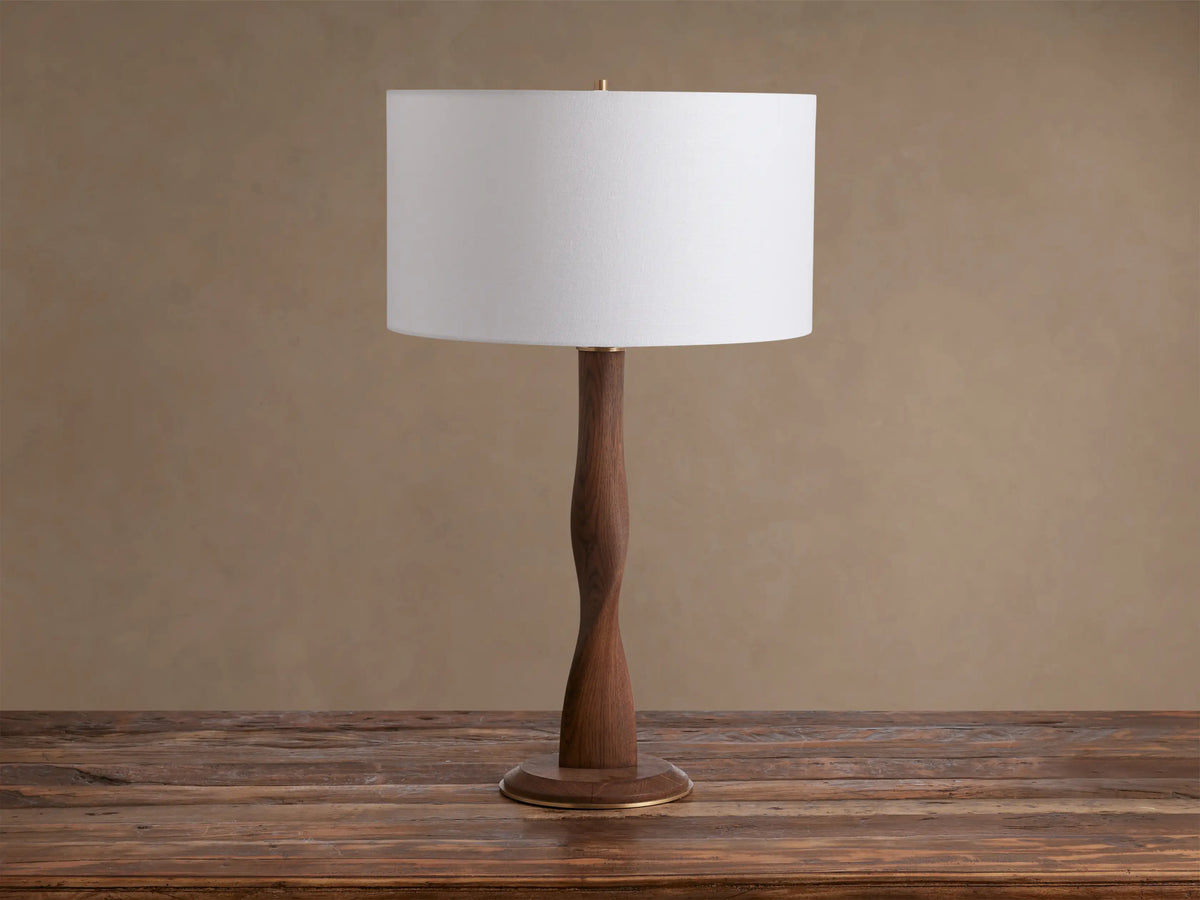 Jonas Table Lamp: White linen shade, curved dark oak wood base with brass accents, on a rustic wooden table.