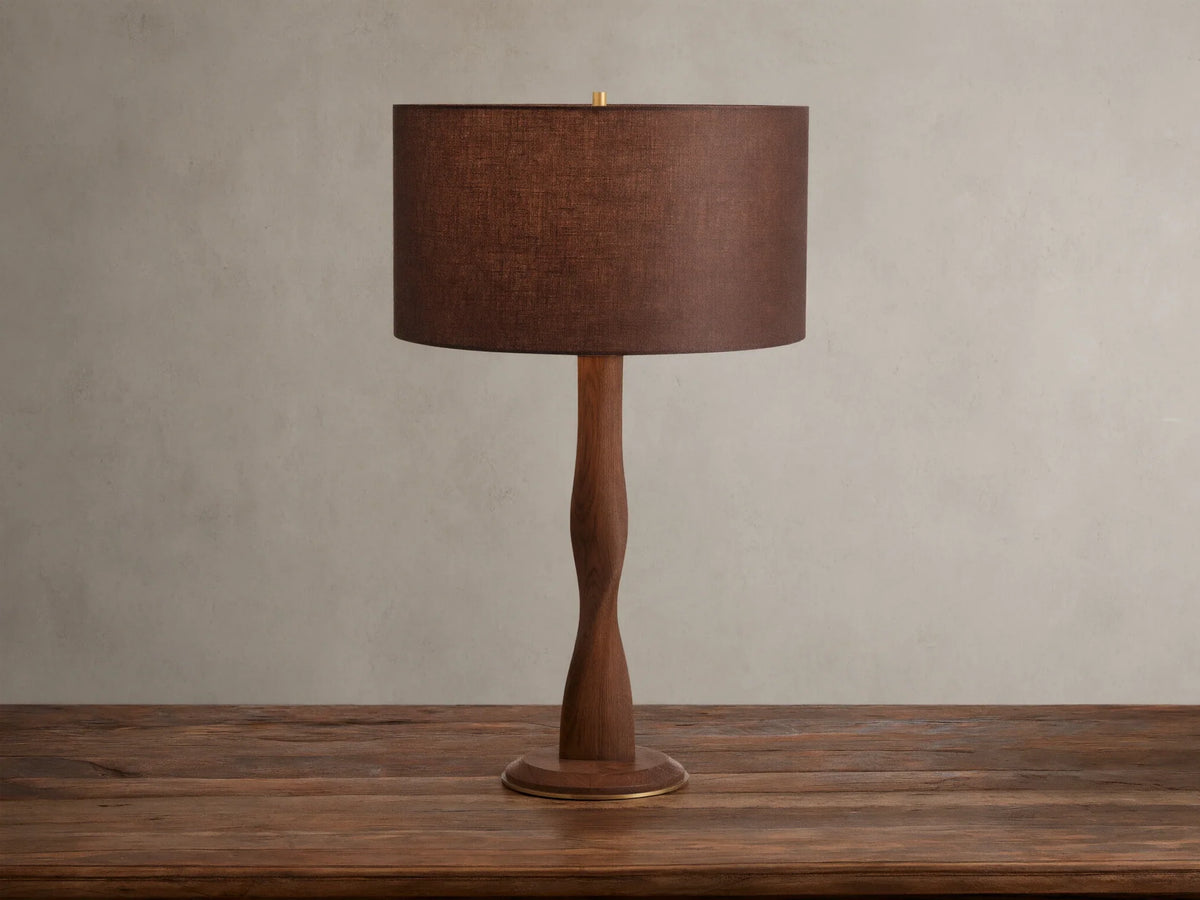 A brown linen shade tops the Jonas Table Lamp, crafted from oak wood with a twisted design and brass accents, on a wooden table.