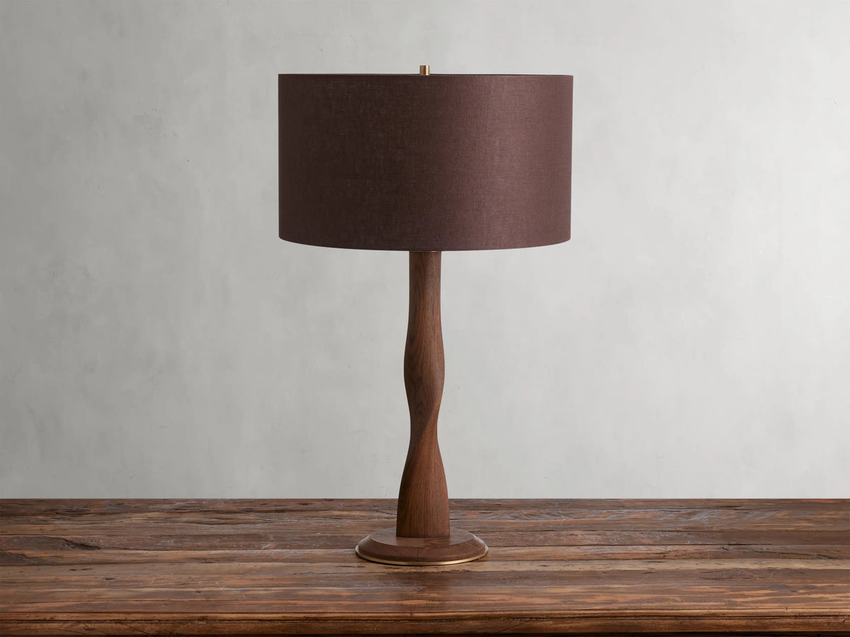 A brown linen shade tops the Jonas Table Lamp, crafted from oak wood with a twisted design and brass accents, on a wooden table.