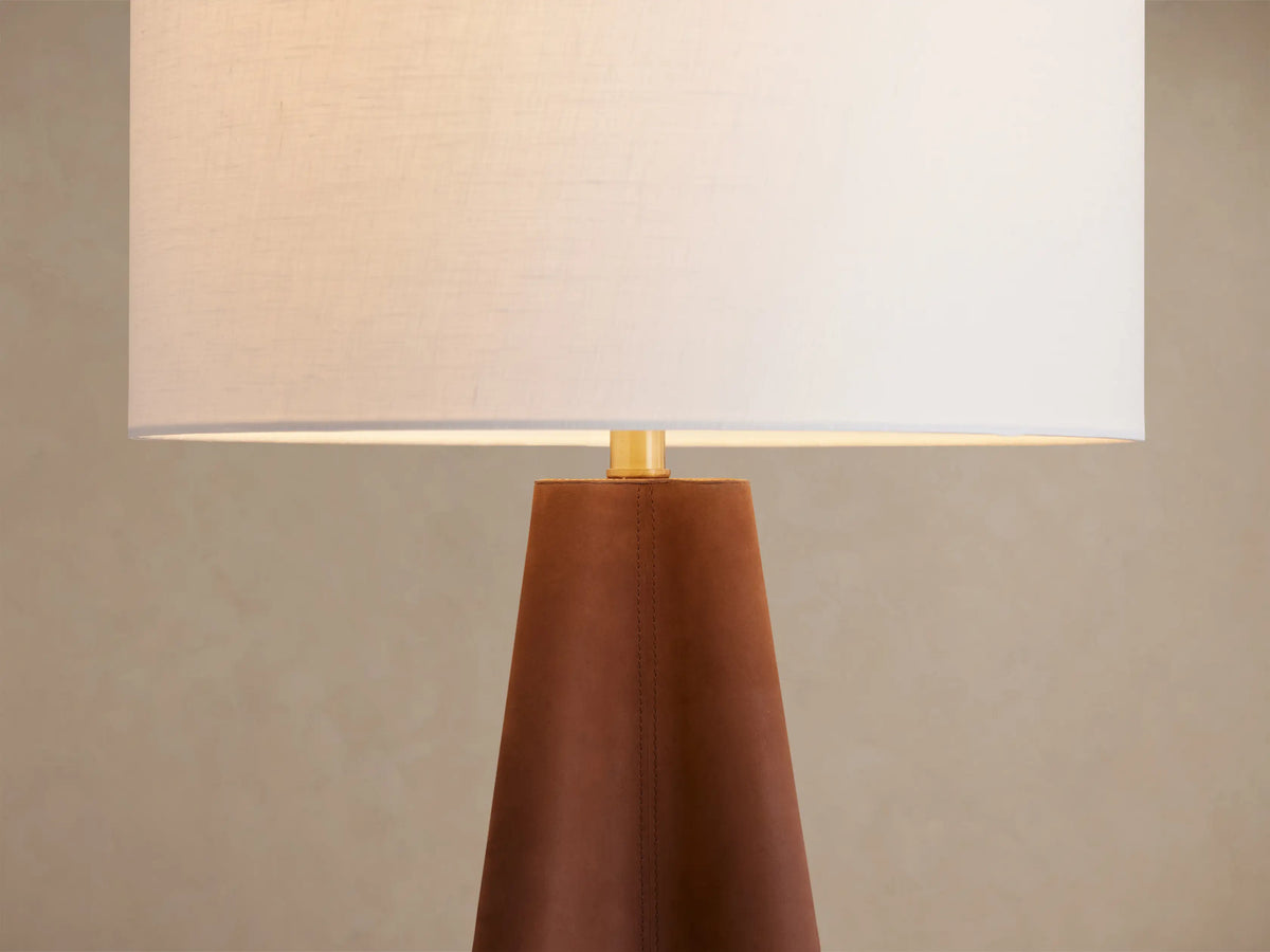 Volant Table Lamp: Tapered dark brown oak wood base with brass accent, topped with a white cylindrical linen shade. On a rustic wood table.