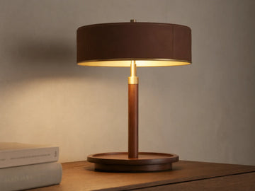 A wooden table lamp with a round dark brown shade, emitting warm light, sits on a wooden surface beside closed books.