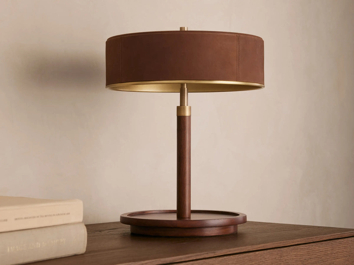 A wooden table lamp with a round dark brown shade, emitting warm light, sits on a wooden surface beside closed books.