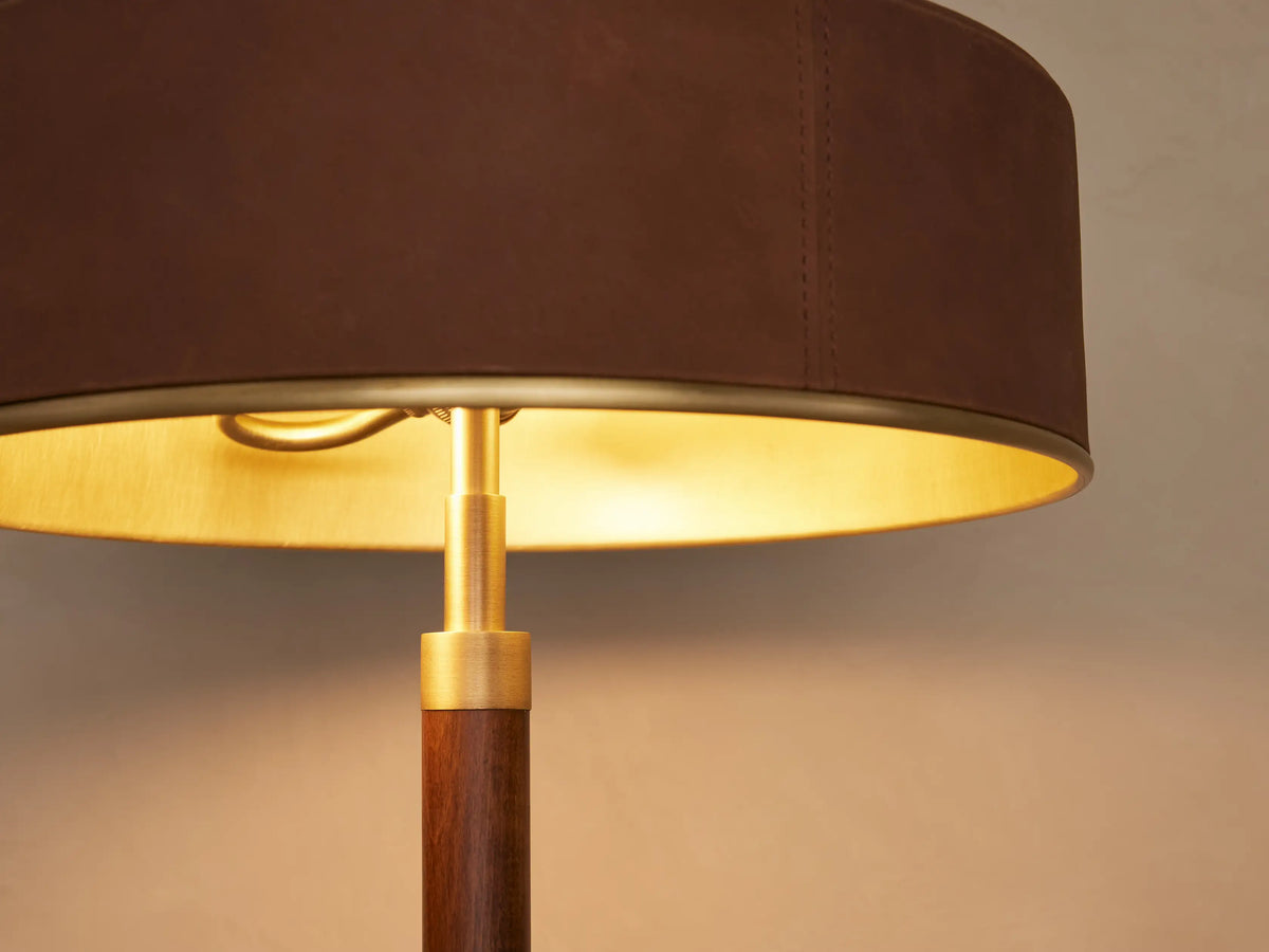 A brown table lamp with a round drum shade and gold interior sits on a wooden surface. The lamp has a sleek cylindrical base.