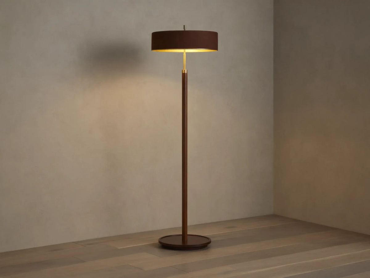 A modern floor lamp with a brown cylindrical shade and slender wooden stand, casting warm light on a wooden floor and neutral wall.