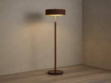 A modern floor lamp with a brown cylindrical shade and slender wooden stand, casting warm light on a wooden floor and neutral wall.
