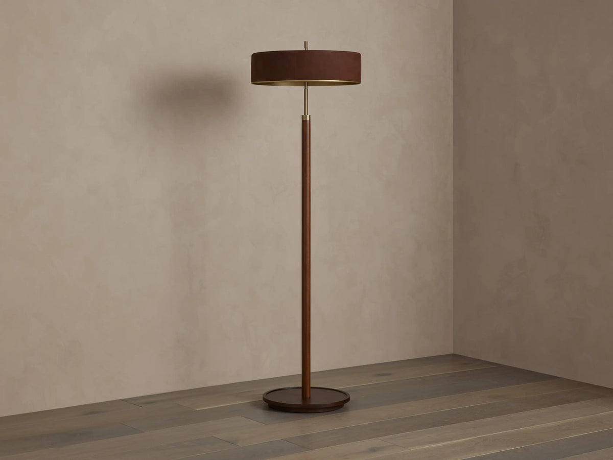 A modern floor lamp with a brown cylindrical shade and slender wooden stand, casting warm light on a wooden floor and neutral wall.