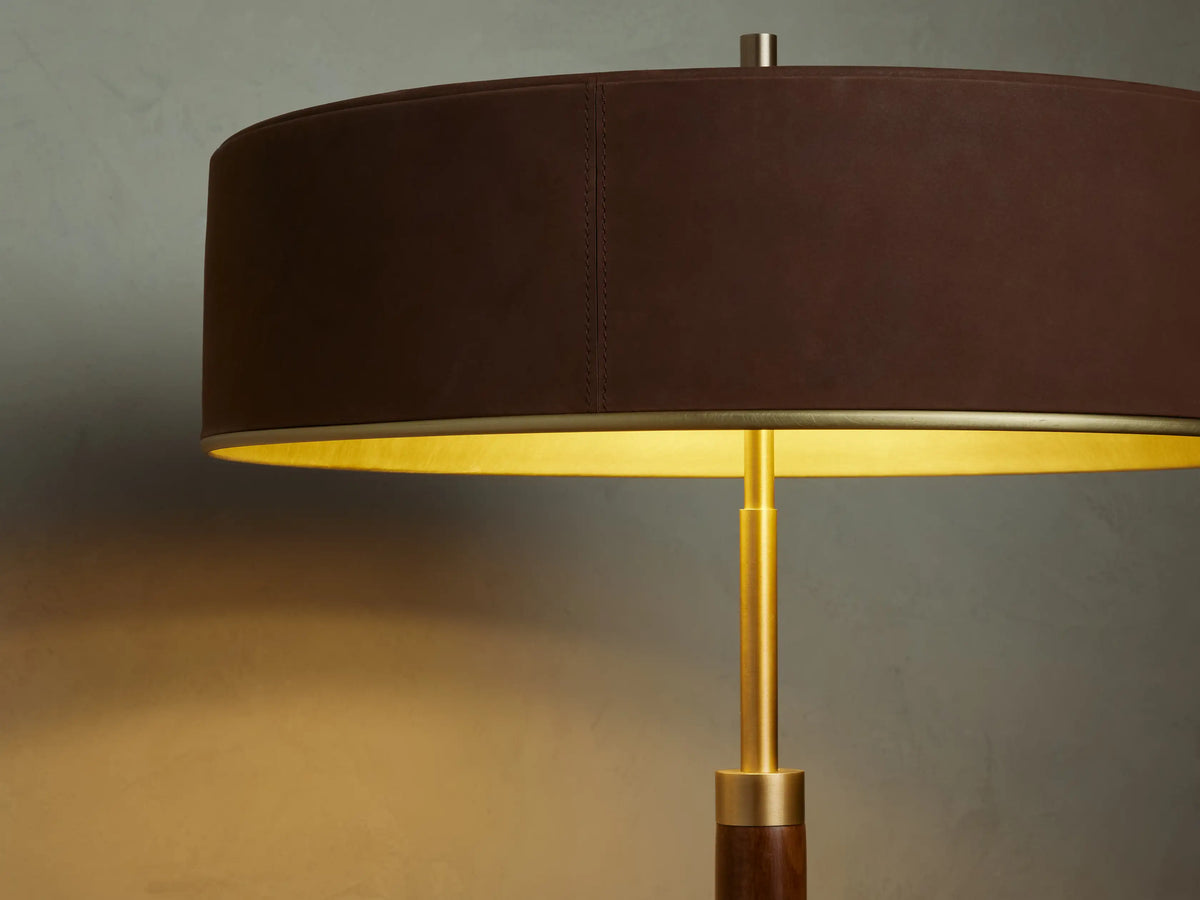 A sleek floor lamp with a dark brown cylindrical shade and base, standing on a wooden floor against a beige wall.
