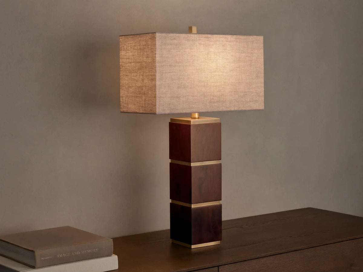 A table lamp with a rectangular fabric shade and a base composed of stacked wooden cubes with gold accents, on a wooden surface.