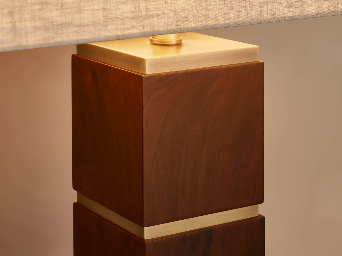 A modern table lamp with a beige rectangular shade and a geometric stacked wood and gold base, placed on a wooden surface.