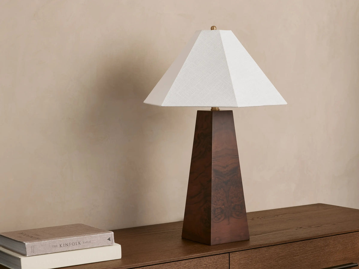A wooden table lamp with a trapezoidal base and a white, pyramid-shaped shade, placed on a wooden surface beside two stacked books.