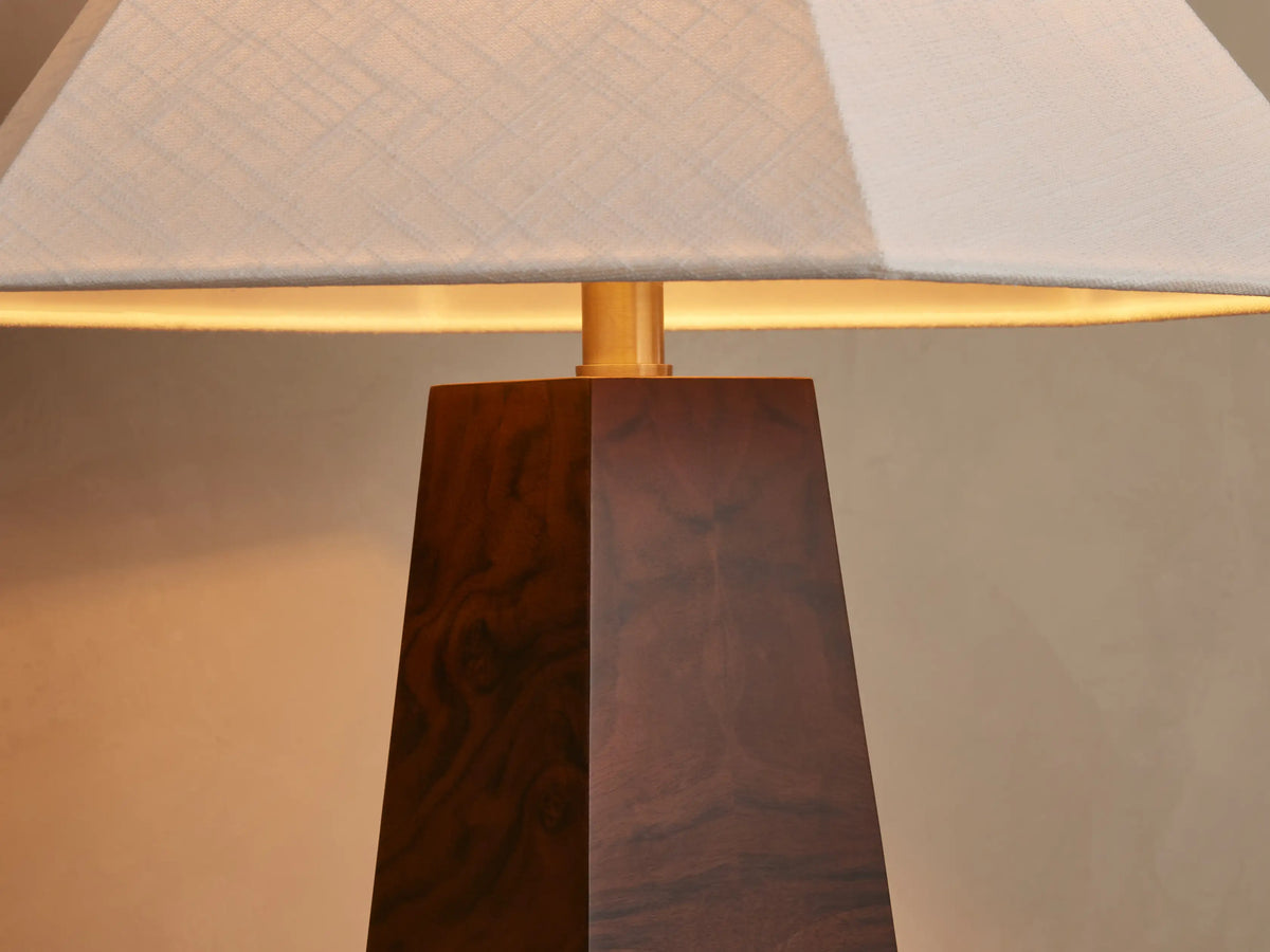 A table lamp with a wooden pyramid-shaped base and a wide, white, tapered square lampshade, placed on a wooden surface next to books.