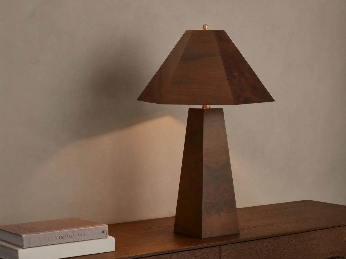 A wooden table lamp with a pyramid-shaped shade and tapered base emits a warm glow on a wooden surface beside stacked books.