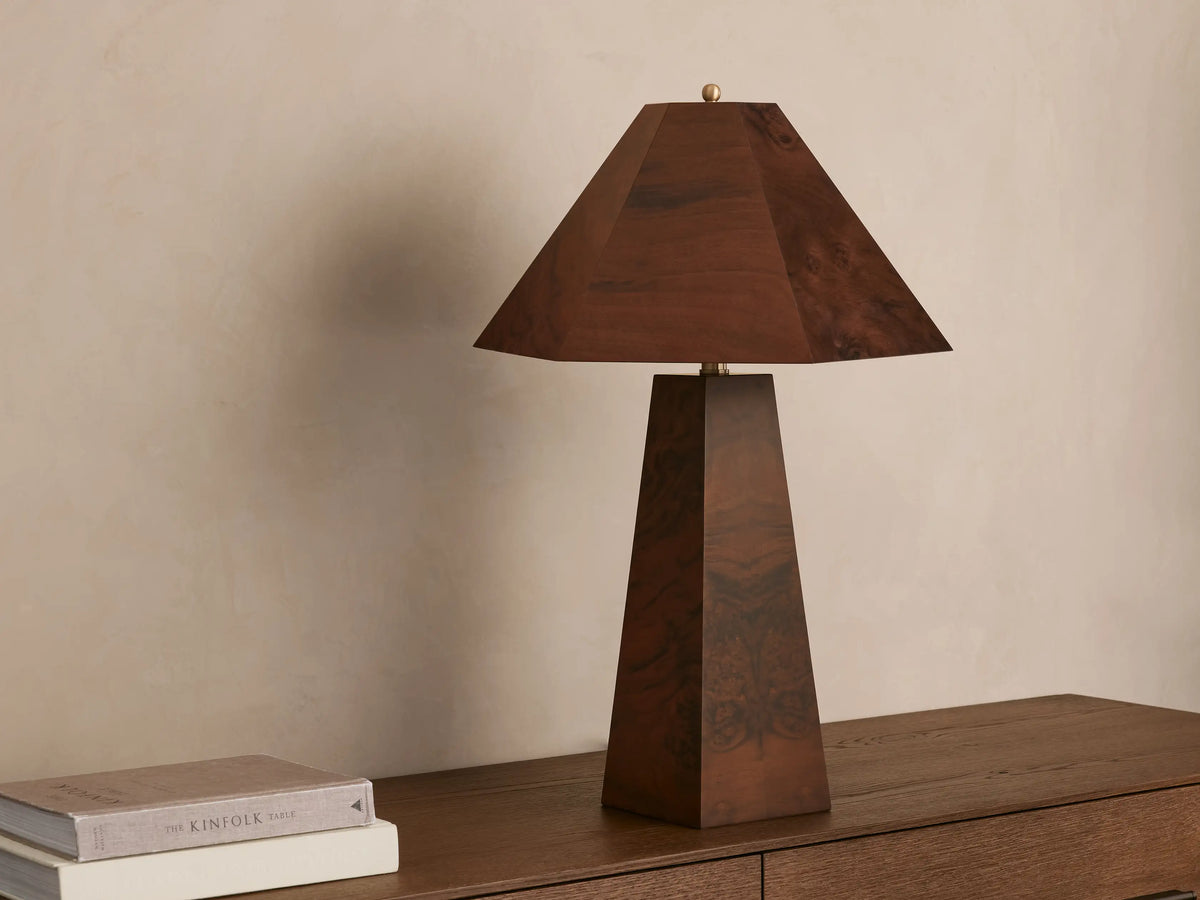 A wooden table lamp with a pyramid-shaped shade and tapered base emits a warm glow on a wooden surface beside stacked books.
