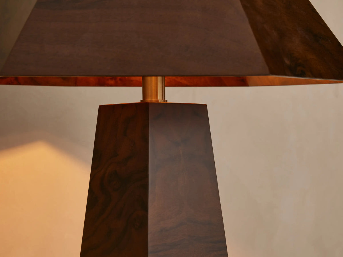 A wooden table lamp with a pyramid-shaped shade and tapered base, crafted from dark wood, sits on a wooden surface by beige wall.