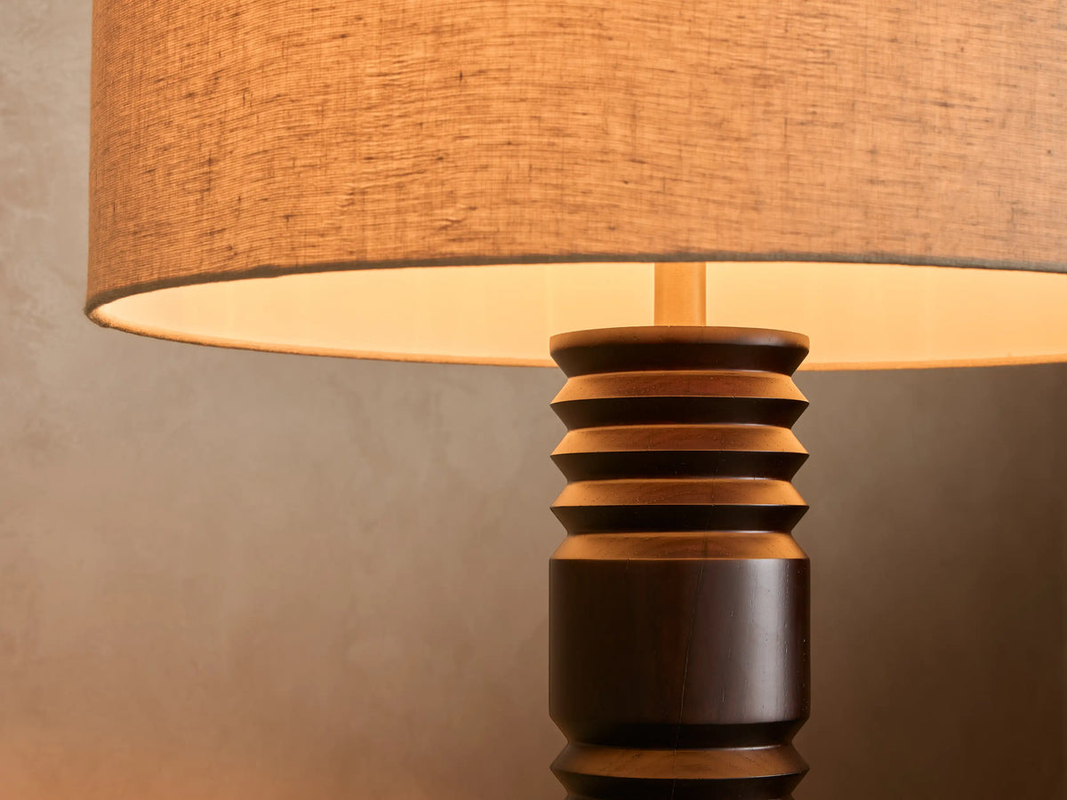 Close-up of a lamp with warm light emanating from a natural fabric shade and a dark brown segmented wooden base.