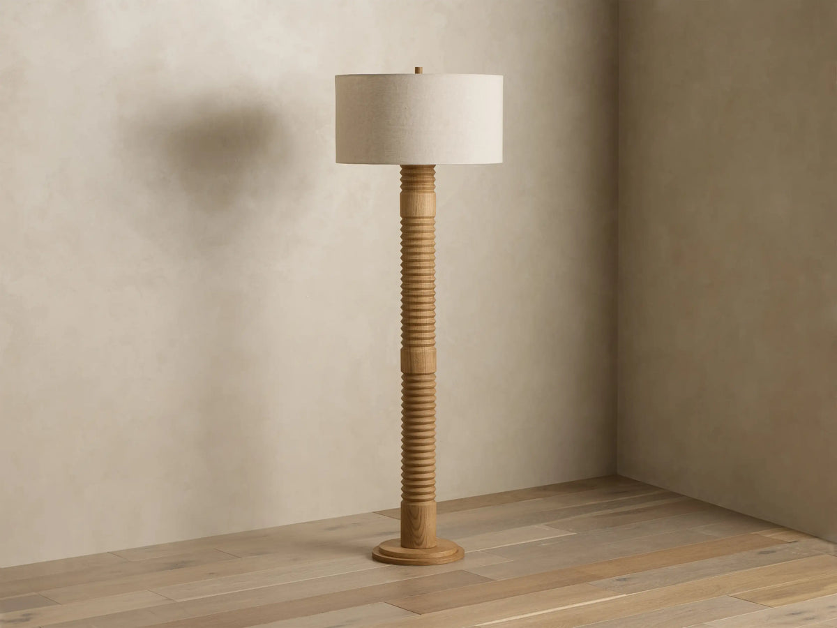 Tall light wood floor lamp with a ribbed stand and glowing beige fabric drum shade, standing in a room corner.