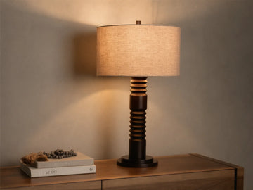 Front view of a dark brown carved base table lamp with a cylindrical light beige fabric shade, illuminated.