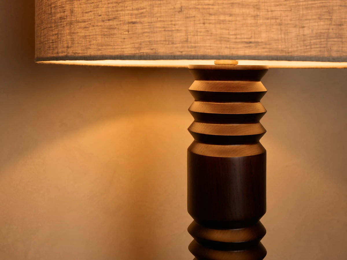 Close-up of a table lamp with a light grey fabric shade emitting a warm glow. The dark brown wood base has a tiered, carved design.