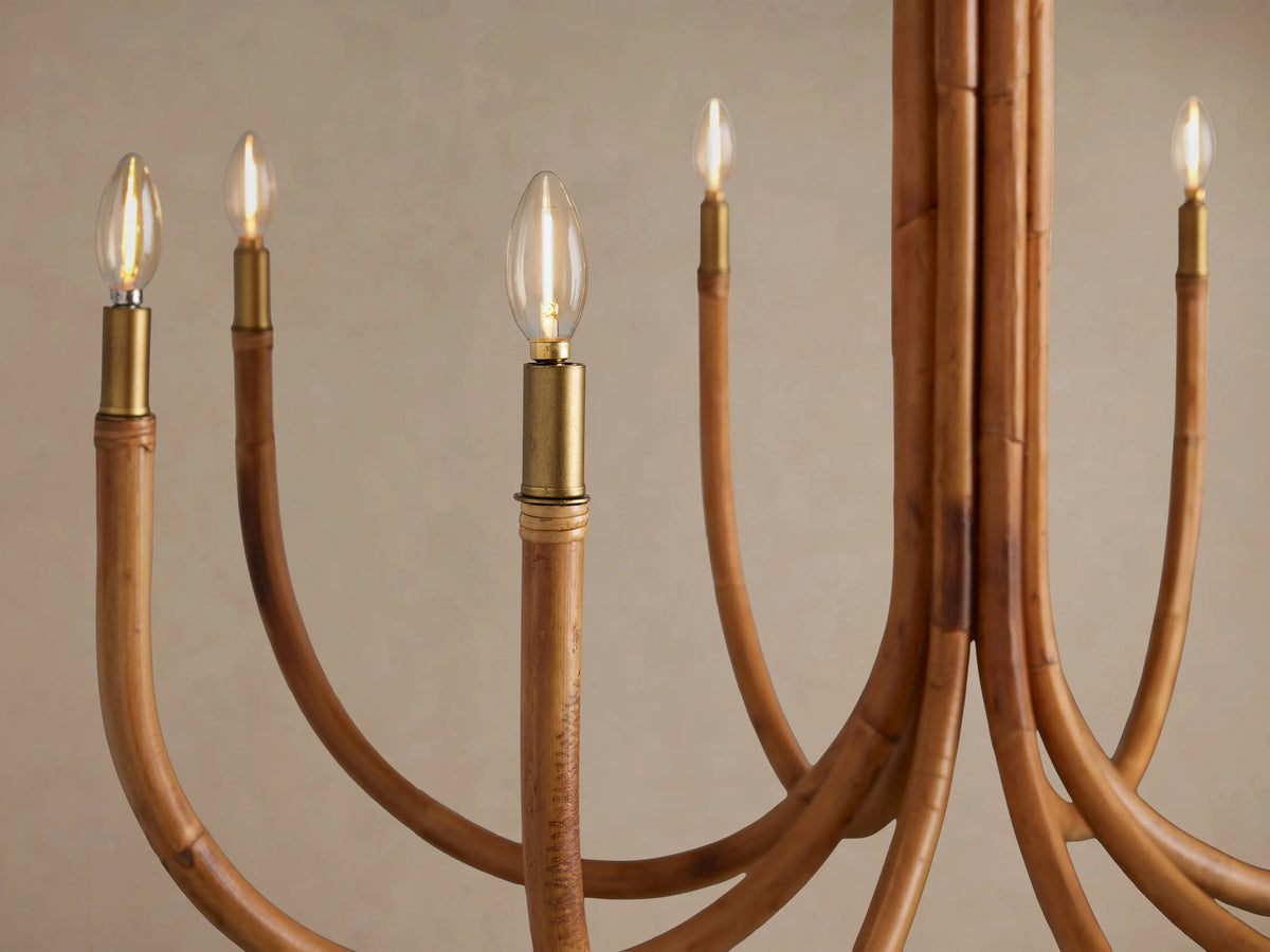 Close-up of the Ravda Round Chandelier: light brown rattan arms with brass accents and glowing candelabra bulbs against a beige wall.