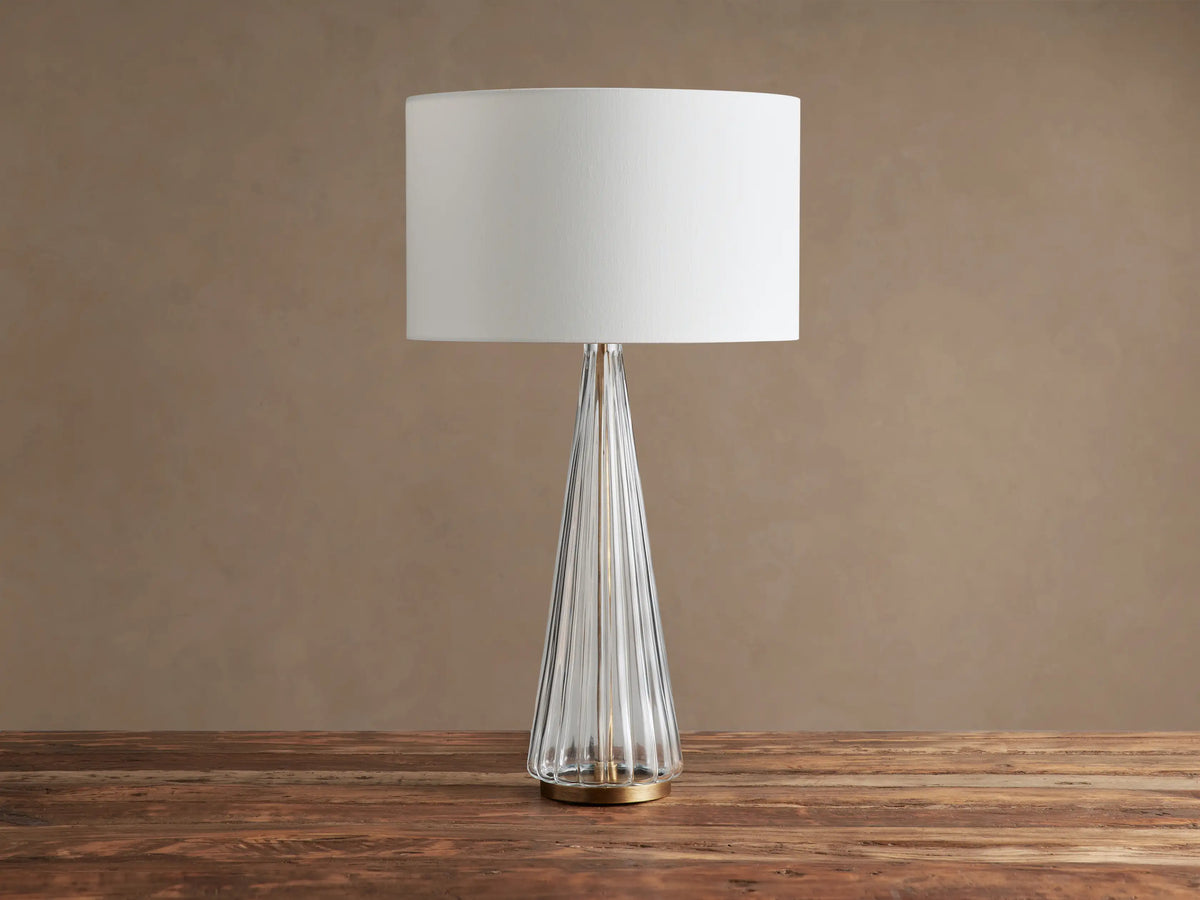 Elisabetta Table Lamp: white linen shade, clear fluted glass conical base, brass finish. On a rustic wood table.