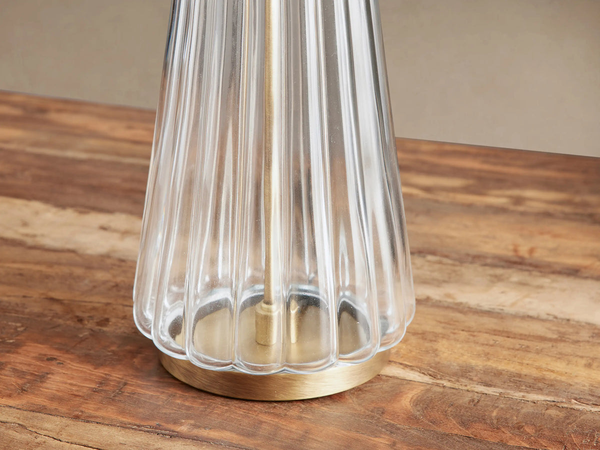Elisabetta Table Lamp with a clear fluted glass tapered base, brass bottom, and white linen shade on a rustic wood table.