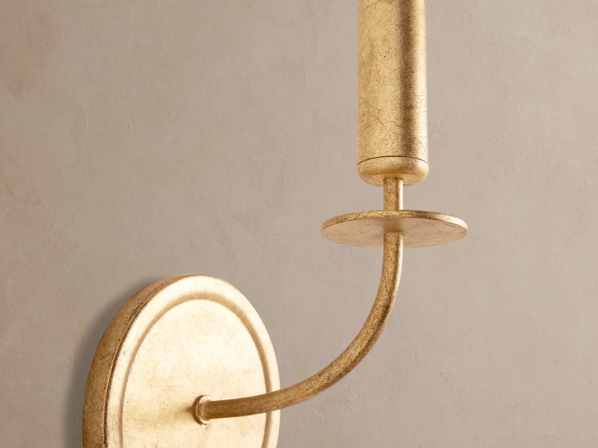 Arnett Sconce: antiqued gold leaf finish wall light. Round backplate, curved arm, and cylindrical bulb holder on a neutral wall.