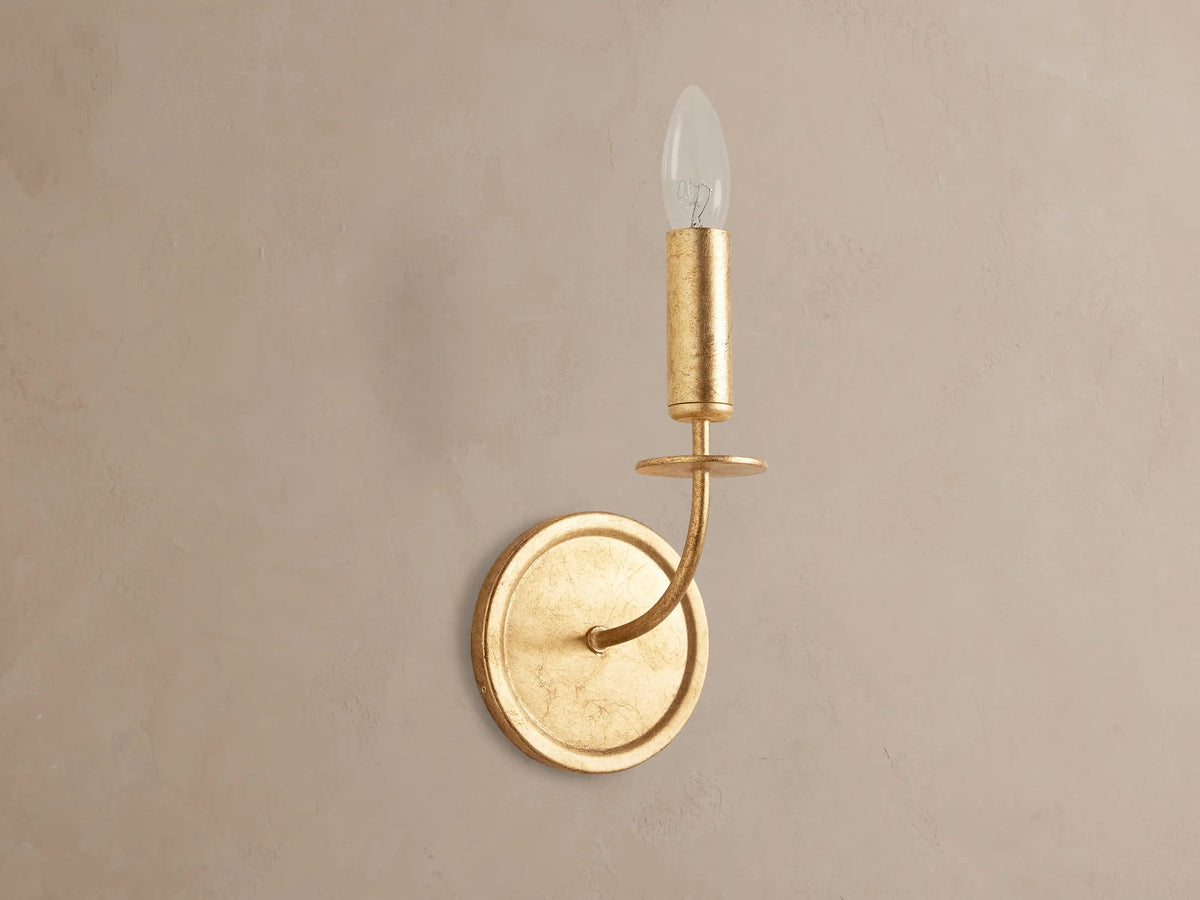 Arnett Sconce: antiqued gold leaf iron sconce with round backplate, curved arm, and candle-style holder with clear bulb.