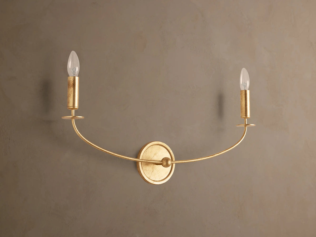 Arnett Double Sconce with an antiqued gold finish, featuring a round backplate and two candle-style lights on a curved arm.