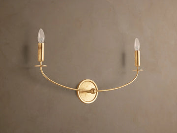Arnett Double Sconce with an antiqued gold finish, featuring a round backplate and two candle-style lights on a curved arm.