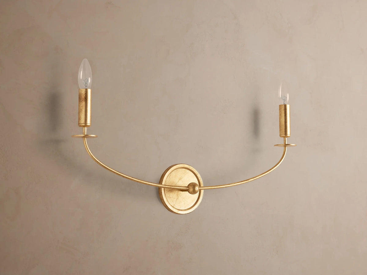 Arnett Double Sconce with an antiqued gold finish, featuring a round backplate and two candle-style lights on a curved arm.