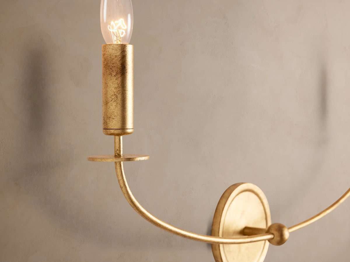 Arnett Double Sconce: antiqued gold iron wall light with two curved arms holding candlestick-style clear bulbs on a textured wall.