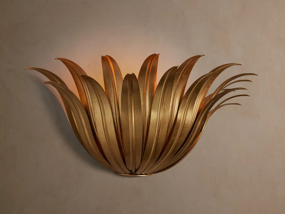 Matilde Sconce, an antique-gold iron wall light resembling layered leaves, casting a warm glow on a neutral wall.