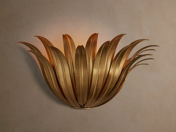 Matilde Sconce, an antique-gold iron wall light resembling layered leaves, casting a warm glow on a neutral wall.