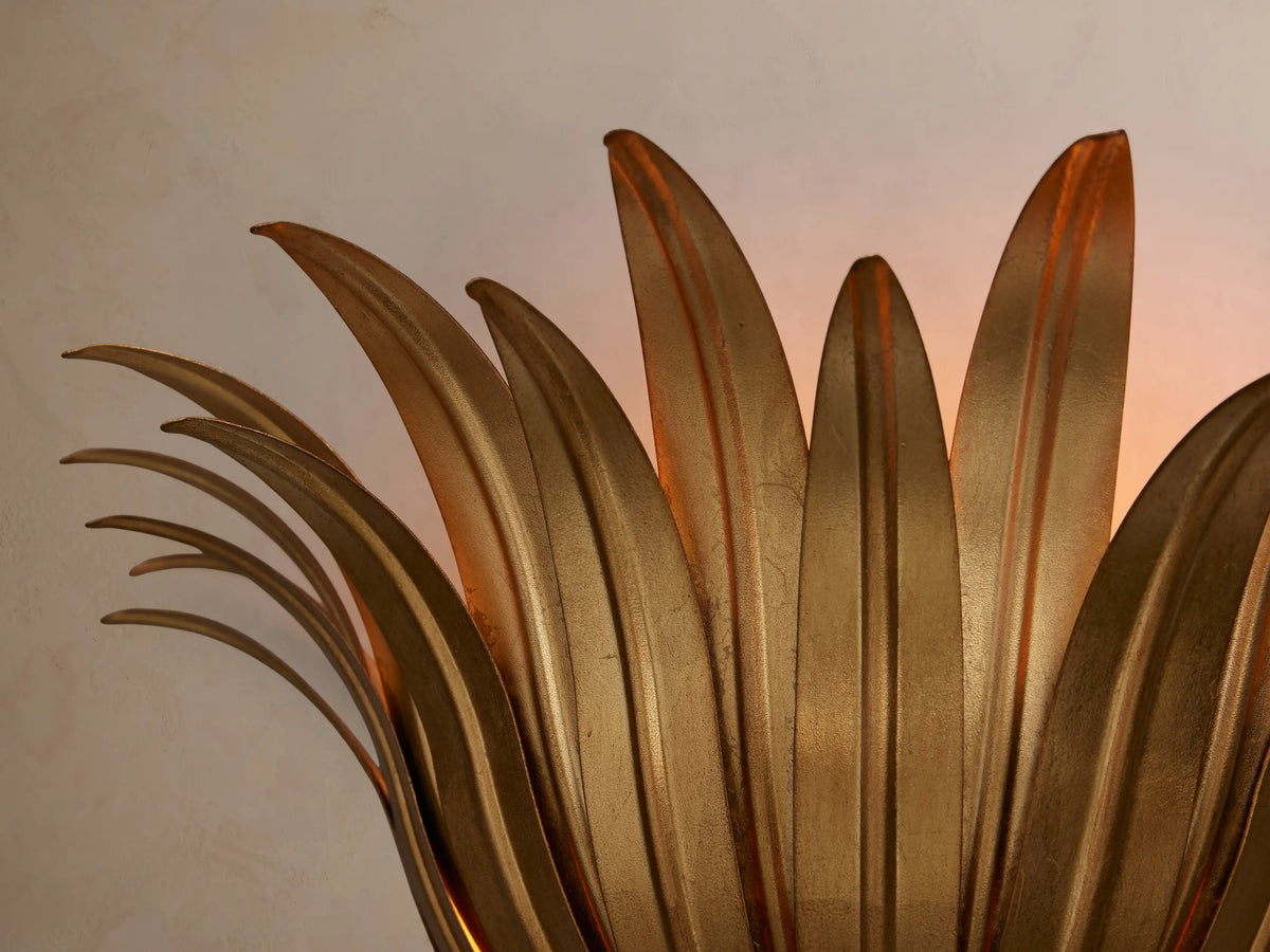 Matilde Sconce, antique-gold iron fixture with fanned leaf design and warm light illuminating a textured wall.