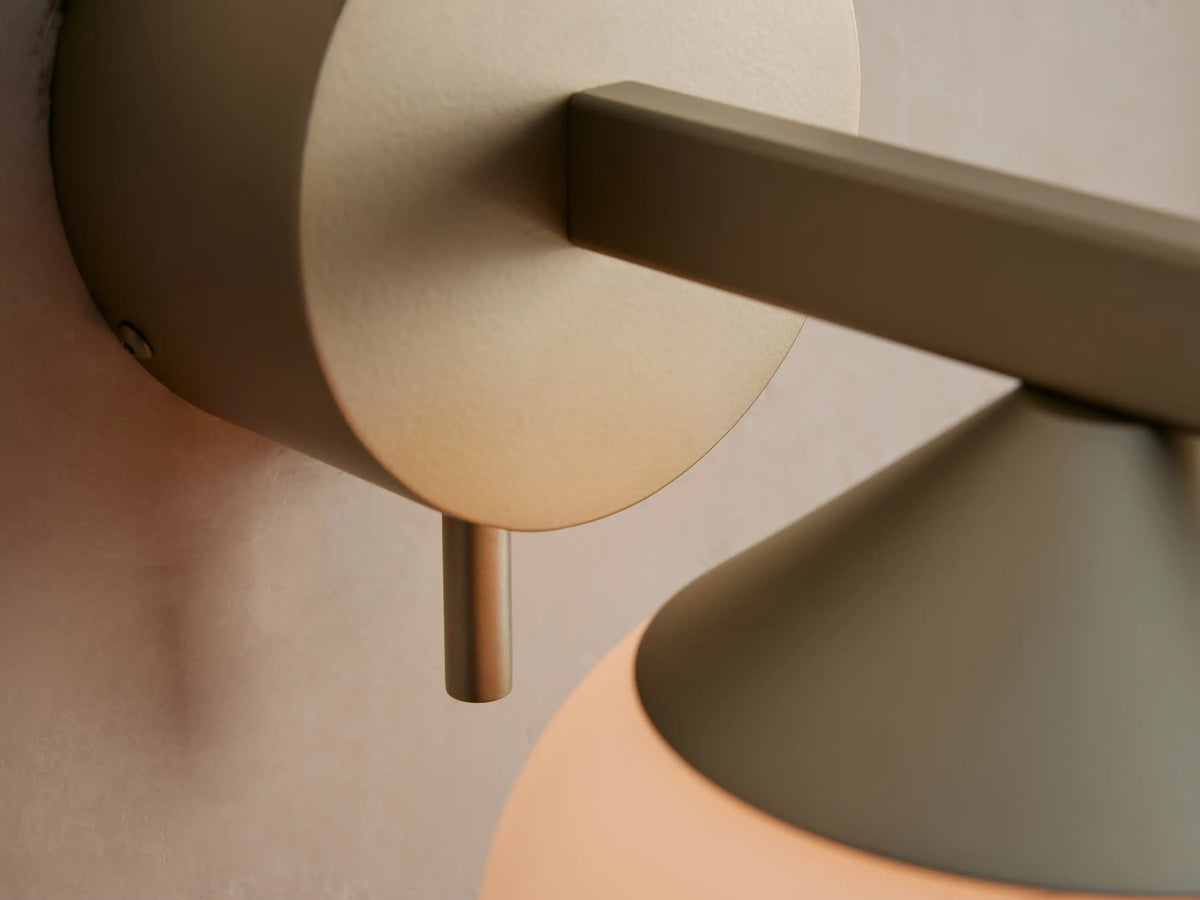 Costella Sconce: champagne/gold iron wall light with round base, angular arm, and spherical white opal glass diffuser on beige wall.