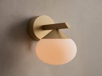 Costella Sconce, a gold iron wall light with a geometric arm and an illuminated, round opal glass diffuser, mounted on a neutral wall.