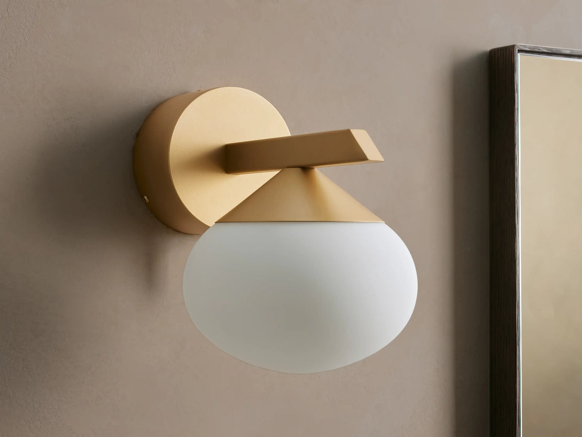 Costella Sconce, a gold iron wall light with a geometric arm and an illuminated, round opal glass diffuser, mounted on a neutral wall.