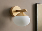 Costella Sconce, a gold iron wall light with a geometric arm and an illuminated, round opal glass diffuser, mounted on a neutral wall.