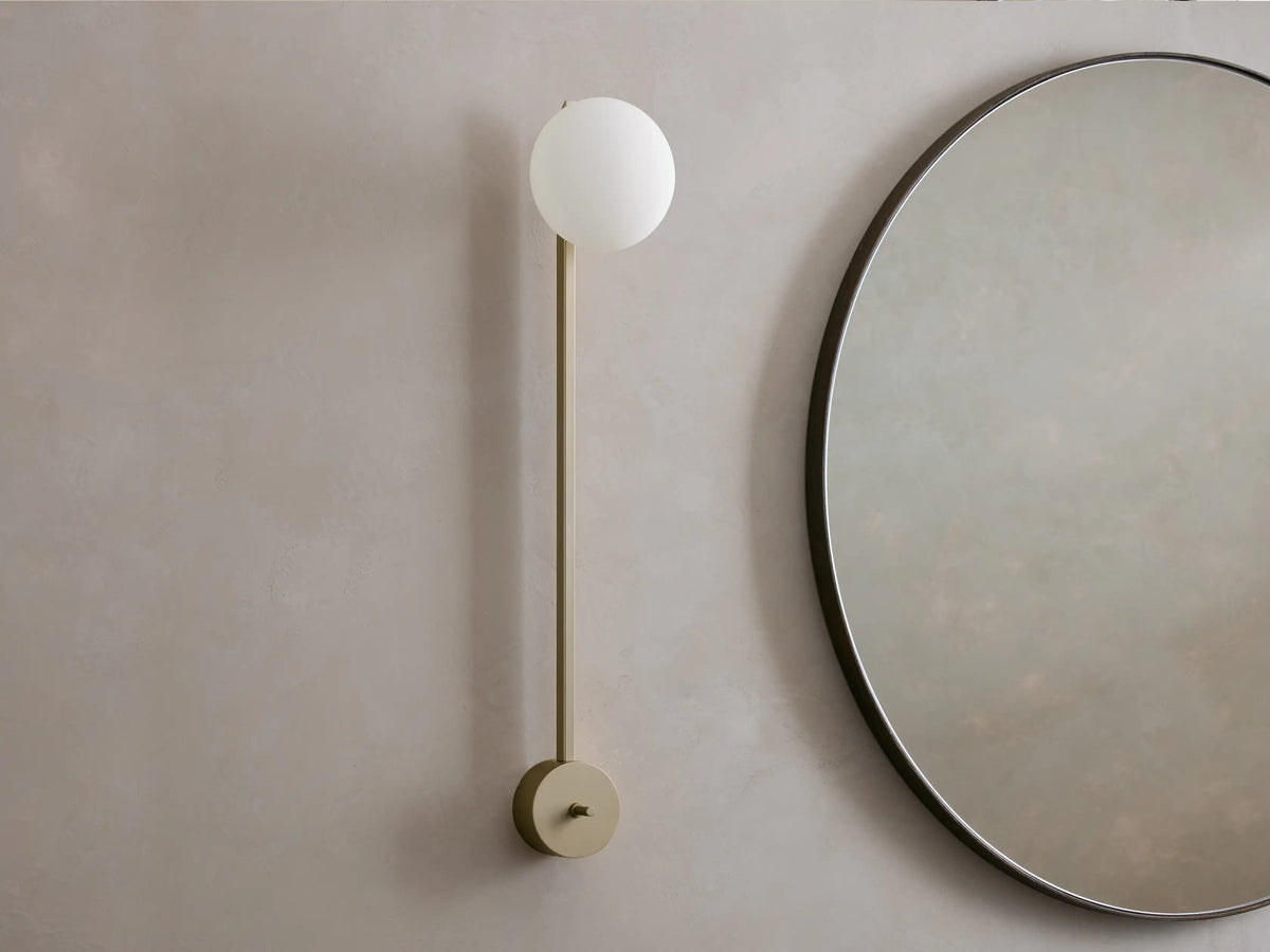 Costella Grand Sconce: a vertical champagne-finished metal bar with a glowing round opal glass diffuser, next to a round mirror.