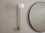 Costella Grand Sconce: a vertical champagne-finished metal bar with a glowing round opal glass diffuser, next to a round mirror.