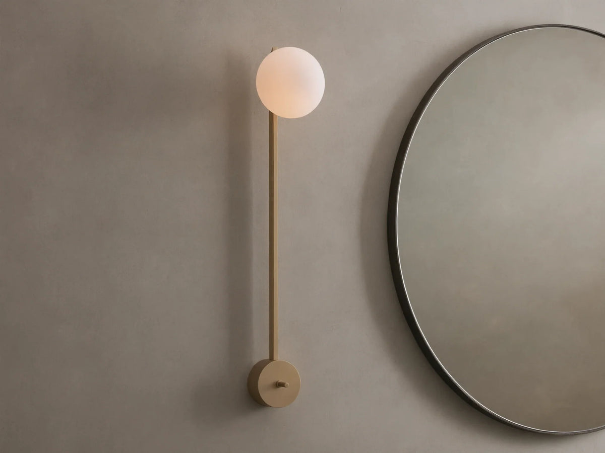 Costella Grand Sconce: a long champagne gold iron wall light with a glowing white spherical opal diffuser, next to a round mirror.