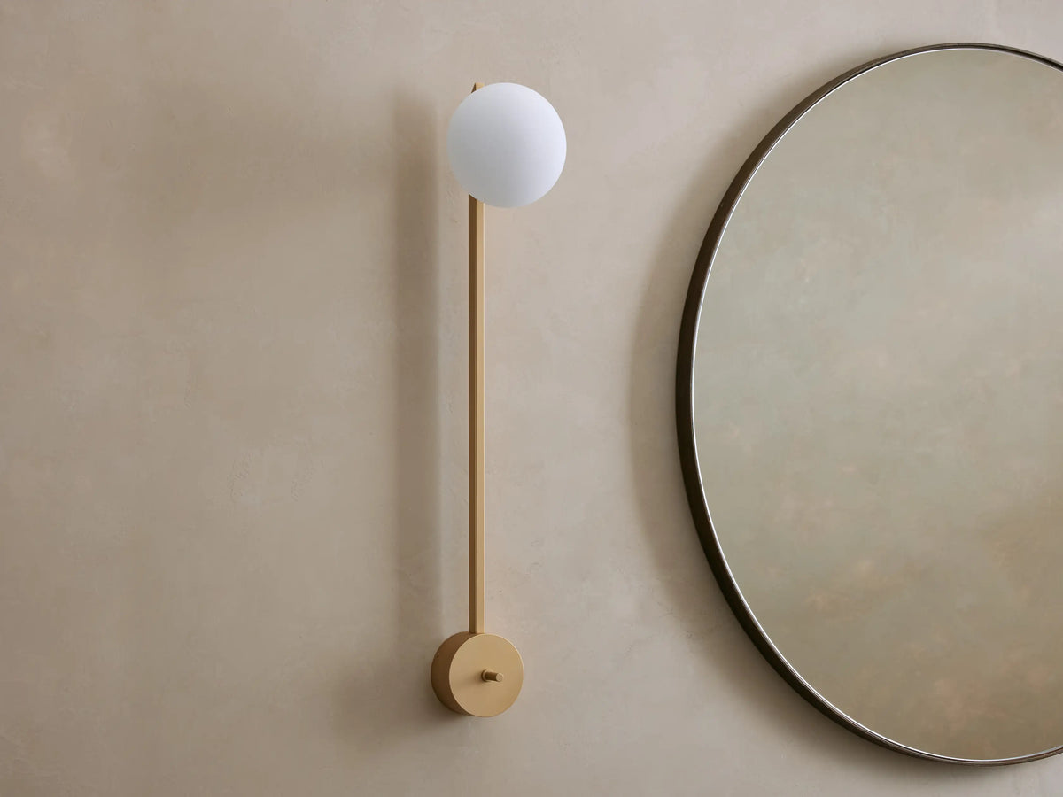 Costella Grand Sconce: a long champagne gold iron wall light with a glowing white spherical opal diffuser, next to a round mirror.