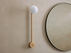 Costella Grand Sconce: a long champagne gold iron wall light with a glowing white spherical opal diffuser, next to a round mirror.