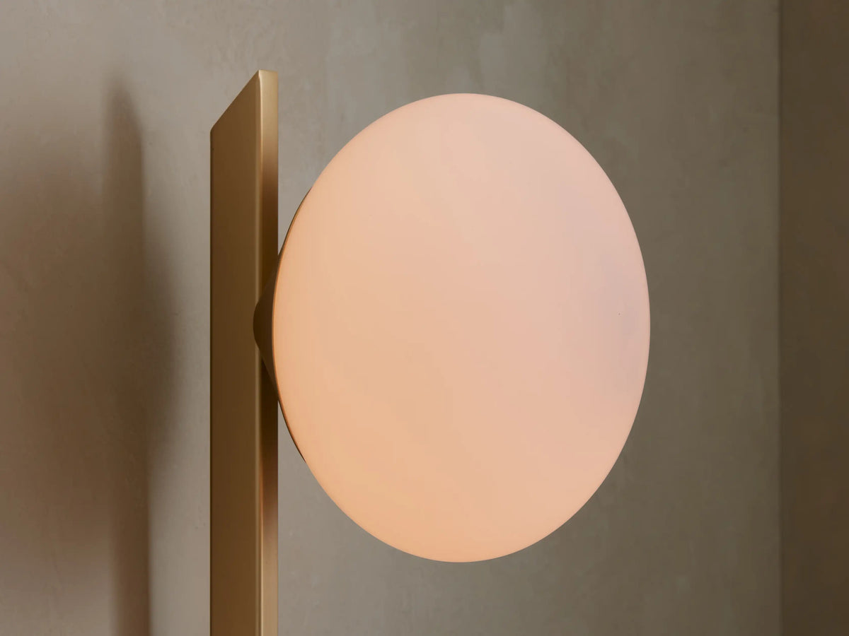 Champagne Costella Grand Sconce: a vertical gold rod with white opal glass globe, on a beige wall next to a round mirror.