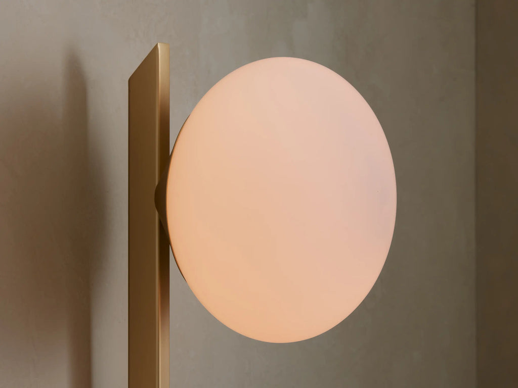 Champagne Costella Grand Sconce: a vertical gold rod with white opal glass globe, on a beige wall next to a round mirror.