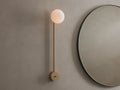 Costella Grand Sconce: a long champagne gold iron wall light with a glowing white spherical opal diffuser, next to a round mirror.