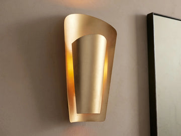 Strato Sconce, a curved, vertical, gold-leafed brass wall sconce with a modern design, illuminated against a neutral wall.