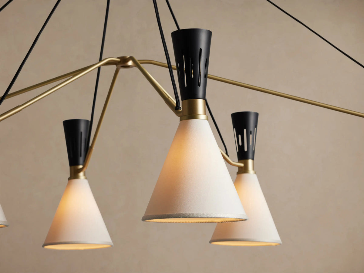 Ramsey Linear Chandelier with six off-white conical shades, matte black accents, and a satin brass frame, hanging against a beige wall.
