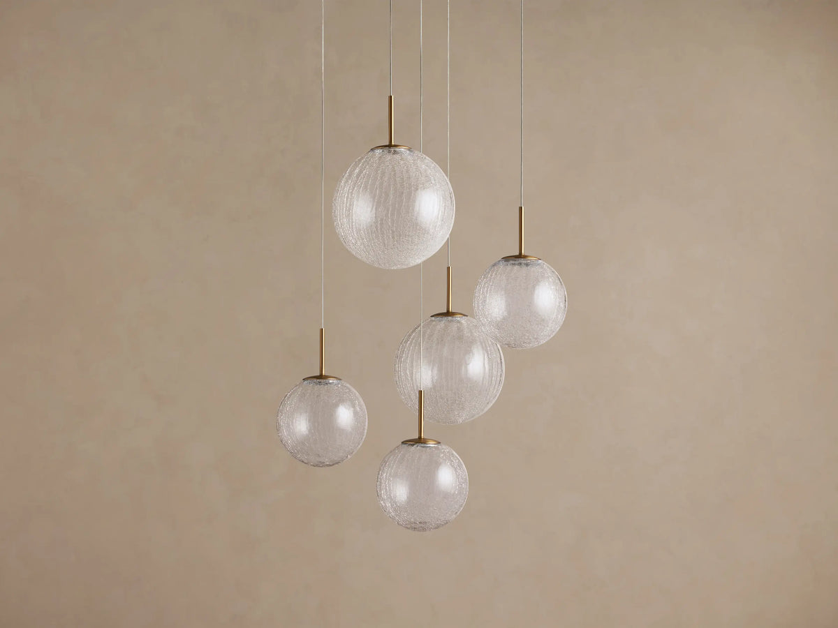 Casen Round Chandelier with five clear crackle glass globes, each suspended by a thin wire and a satin brass stem, against a beige wall.
